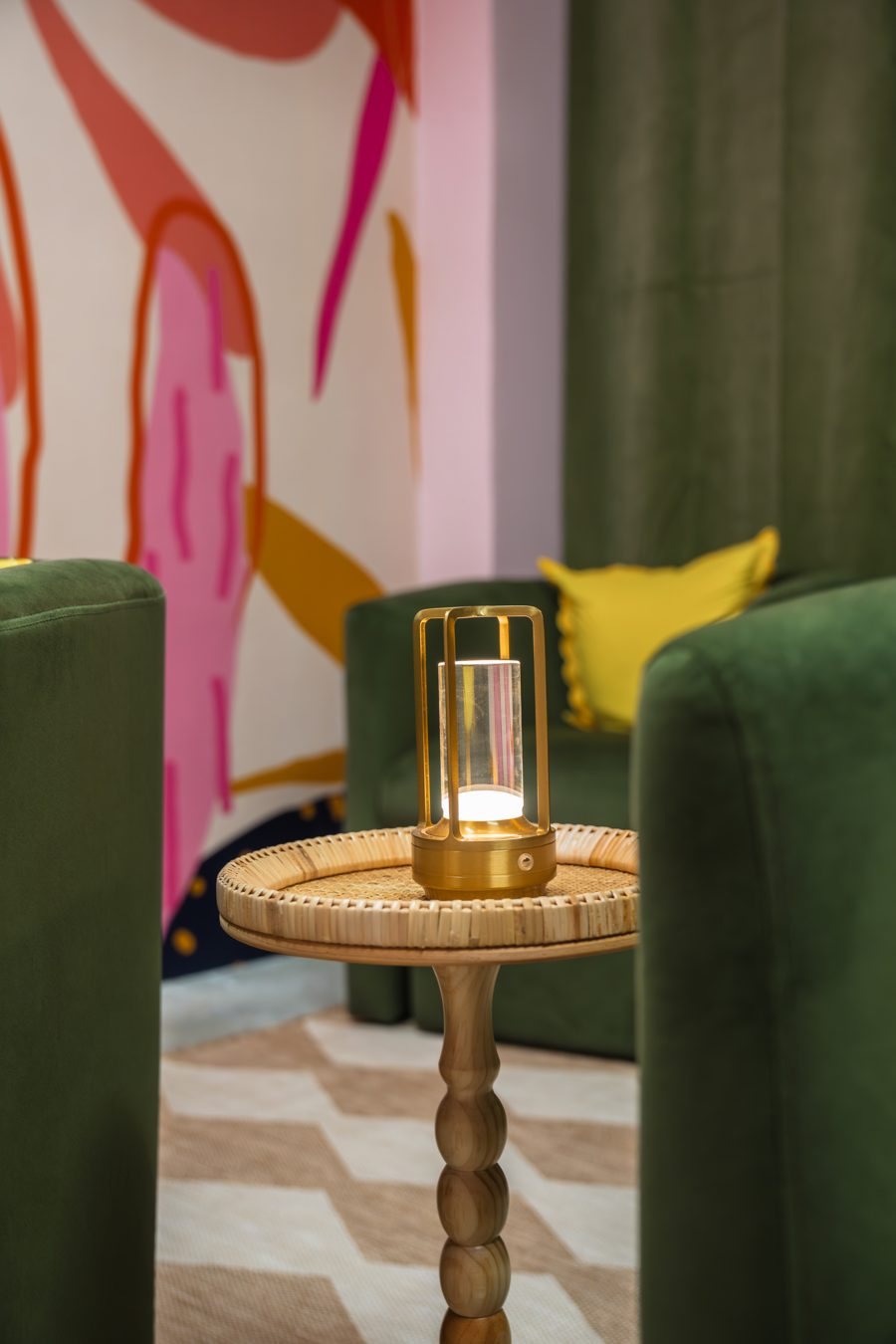 Gold lantern on a rattan side table between green velvet chairs